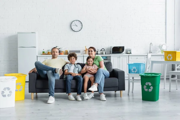 Glückliche Familie sitzt auf Couch in der Nähe von Mülltonnen und Boxen mit Recyclingschild in der Küche — Stockfoto