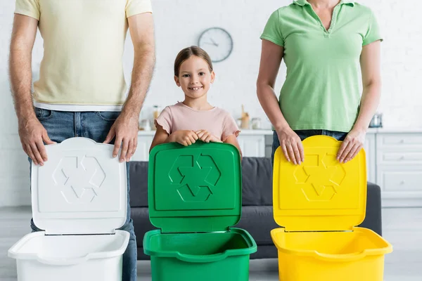 Lächelndes Mädchen blickt in die Kamera in der Nähe von Eltern und Mülltonnen mit Recyclingschild — Stockfoto