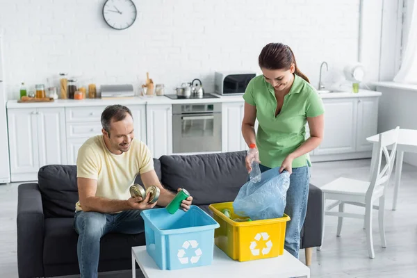 Femme heureuse mettant la bouteille dans un sac poubelle près du mari et des boîtes avec panneau de recyclage à la maison — Photo de stock