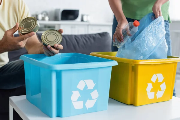 Vista recortada del hombre sosteniendo latas de lata cerca de esposa y cajas con cartel de reciclaje en la cocina - foto de stock