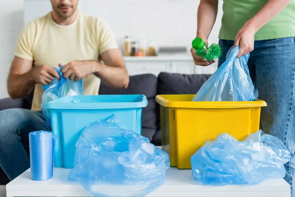 Vue recadrée de la femme tenant sac poubelle et bouteille près des boîtes et mari flou dans la cuisine — Photo de stock