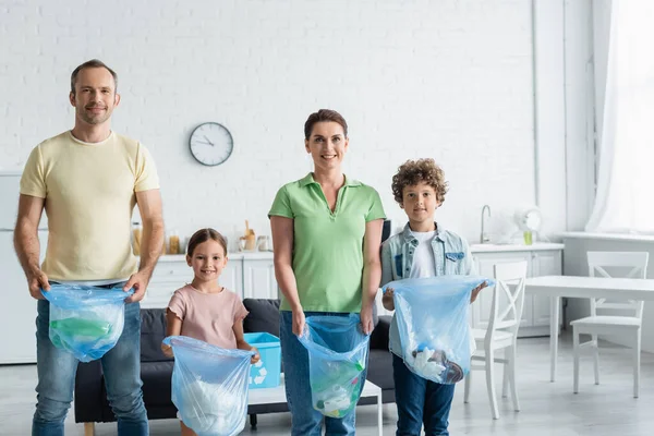 Smiling family holding trash bags and looking at camera in kitchen — Stock Photo