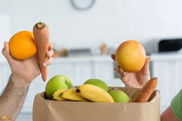 Cropped view of couple holding fruits near paper bag in kitchen — Stock Photo