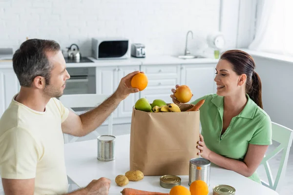 Улыбающаяся пара берет фрукты из бумажного пакета на кухне — стоковое фото