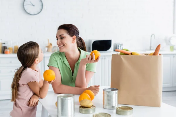 Mère et enfant souriants tenant des oranges près de la nourriture et du papier artisanal dans la cuisine — Photo de stock