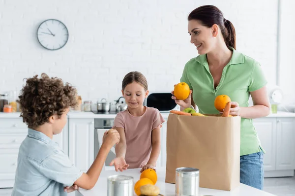 Femme souriante tenant des oranges fraîches près du sac en papier et des enfants dans la cuisine — Photo de stock