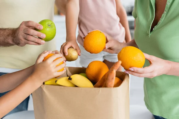 Cropped view of kids and parents holding fruits near craft bag — Stock Photo