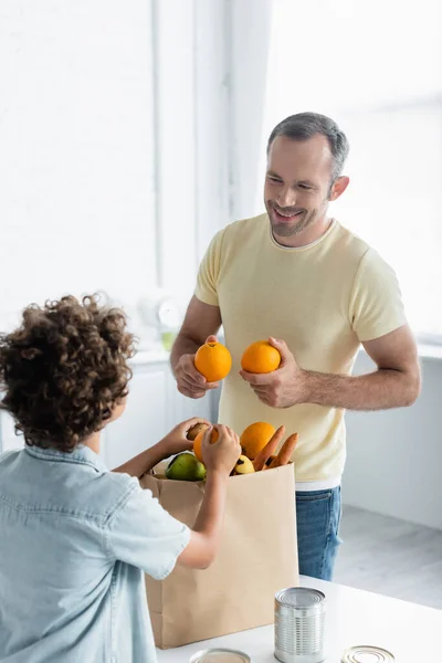 Padre sorridente che tiene le arance vicino al figlio sfocato e al sacchetto di carta in cucina — Foto stock