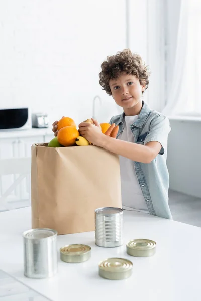 Garçon souriant tenant de la nourriture près du sac en papier et boîtes de conserve dans la cuisine — Photo de stock