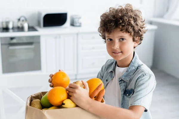 Niño positivo sosteniendo frutas cerca de la bolsa de artesanía en la cocina - foto de stock