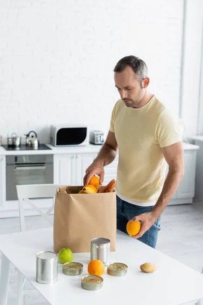 Hombre tomando frutas de la bolsa de papel en la mesa en la cocina - foto de stock