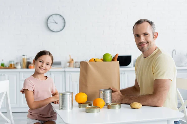 Positivo padre e figlia guardando la fotocamera vicino al sacchetto di carta e cibo in cucina — Foto stock