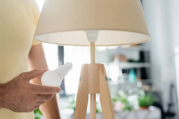 Vista recortada del hombre sosteniendo la bombilla de ahorro de energía cerca de la lámpara de pie en casa - foto de stock