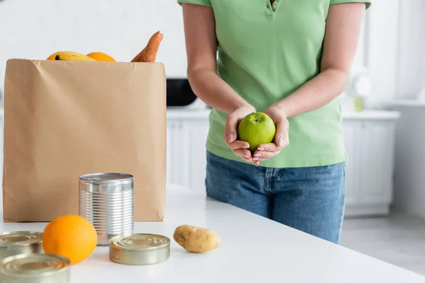 Ausgeschnittene Ansicht einer Frau mit Apfel in der Nähe von Lebensmitteln und Papiertüten in der Küche — Stockfoto