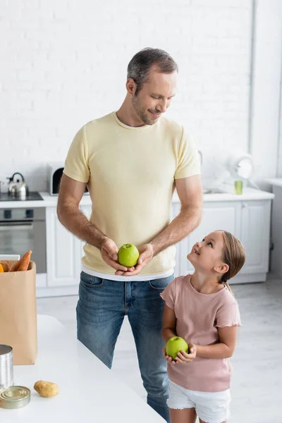 Sonriente padre e hija sosteniendo manzanas cerca de la bolsa de papel en la cocina - foto de stock