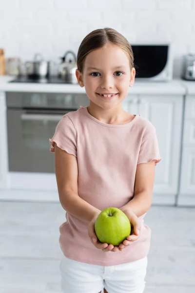 Улыбающаяся девушка держит свежее яблоко и смотрит в камеру на кухне — стоковое фото