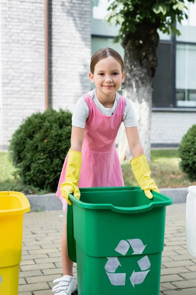 Smiling kid in rubber gloves standing near trash bin with recycle sign outdoors — Stock Photo