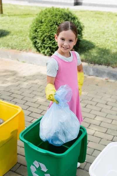 Smiling child in rubber gloves holding trash bag near can with recycle sign on urban street — Stock Photo