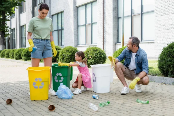 Família com filha em luvas de borracha classificando lixo em latas com sinal de reciclagem ao ar livre — Fotografia de Stock