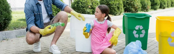 Chica sonriente en guantes de goma sosteniendo basura cerca de padre y latas con cartel de reciclaje en la calle urbana, pancarta - foto de stock