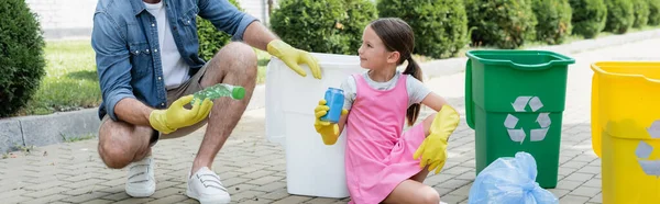 Ragazza sorridente in guanti di gomma che tiene spazzatura vicino al padre e lattine con cartello di riciclo sulla strada urbana, banner — Foto stock