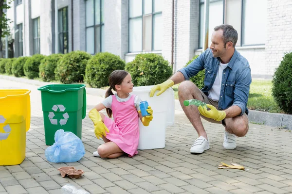 Figlia sorridente e padre in guanti di gomma che tengono cestino vicino a lattine con simbolo di riciclaggio all'aperto — Foto stock