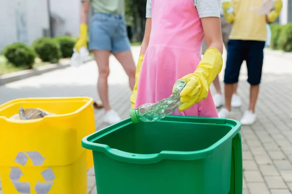 Vista recortada de chica en guantes de goma sosteniendo botella cerca de bote de basura al aire libre - foto de stock