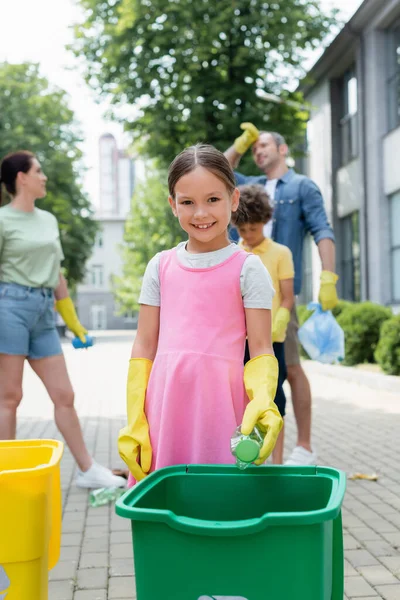 Lächelndes Kind in Gummihandschuhen, das in die Kamera blickt, während es Mülleimer in der Nähe hält und Familie im Freien begräbt — Stockfoto