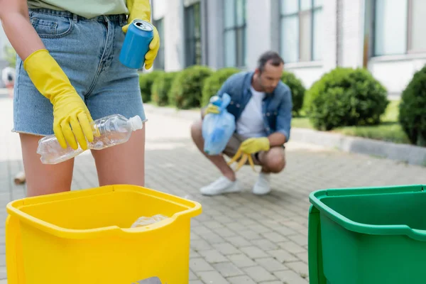 Cropped view of woman holding trash near can and blurred husband outdoors — Stock Photo