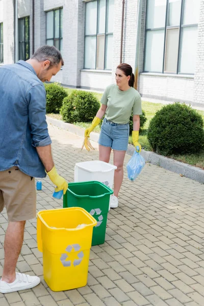 Жінка тримає сміття біля чоловіка і сміттєвих баків з символом переробки на відкритому повітрі — Stock Photo