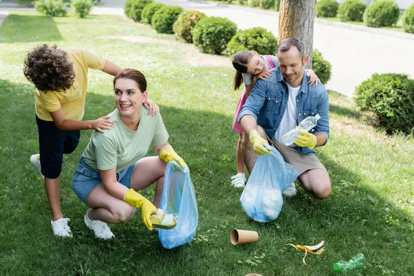 Bambini allegri che abbracciano i genitori con sacchi della spazzatura sul prato — Foto stock