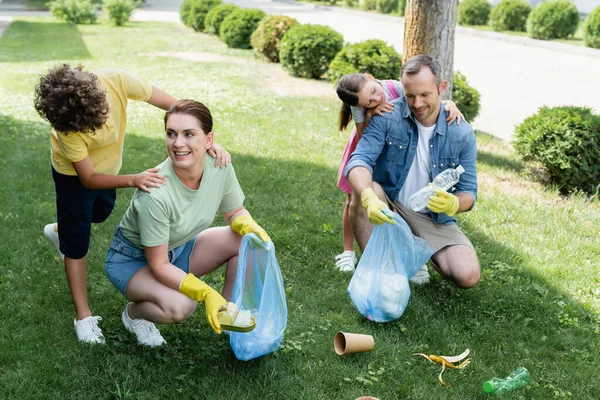 Niños alegres abrazando a los padres con bolsas de basura en el césped - foto de stock