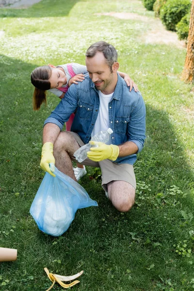 Hija abrazando padre con bolsa de basura en el césped - foto de stock