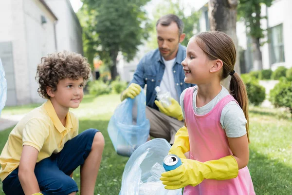 Enfant heureux tenant boîte d'étain et sac poubelle près flou frère et papa à l'extérieur — Photo de stock