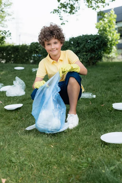 Enfant souriant à la caméra tout en tenant sac poubelle près des ordures sur la pelouse — Photo de stock