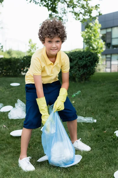 Ragazzo positivo in guanti di gomma sacchetto di tenuta vicino spazzatura sull'erba — Foto stock