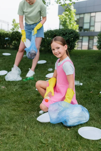 Smiling girl looking at camera while blurred mother collecting trash outdoors — Stock Photo