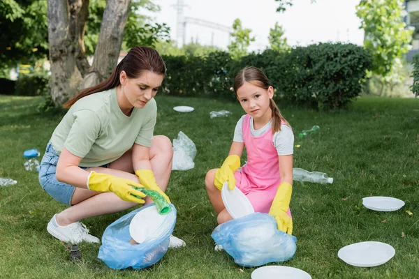 Chica mirando a la cámara cerca de la madre con bolsa de basura en la hierba - foto de stock