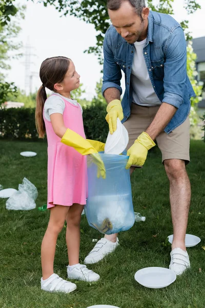Sonriente chica sosteniendo bolsa cerca de padre con basura al aire libre - foto de stock