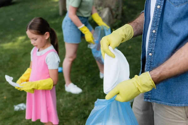 Hombre con guantes de goma poniendo basura en la bolsa cerca borrosa hija y esposa al aire libre - foto de stock