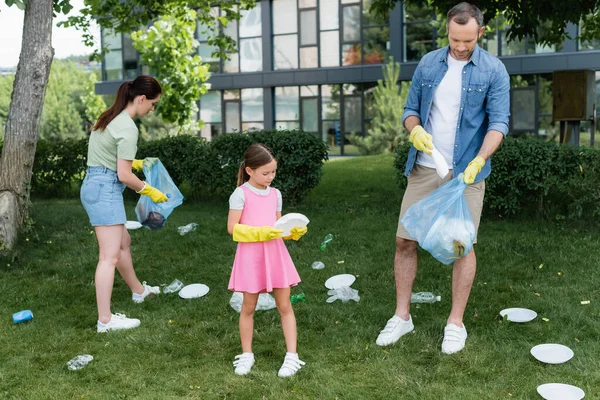 Niño sosteniendo basura plástica cerca de padres con bolsas de basura al aire libre - foto de stock
