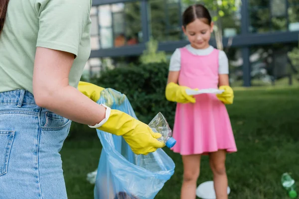 Femme en gants de caoutchouc mettre bouteille dans un sac poubelle près de l'enfant flou à l'extérieur — Photo de stock