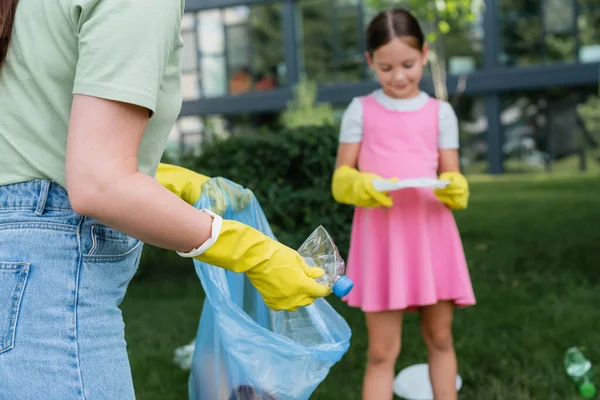 Frau in Gummihandschuhen steckt Flasche in Müllsack neben verschwommenem Kind im Freien — Stockfoto