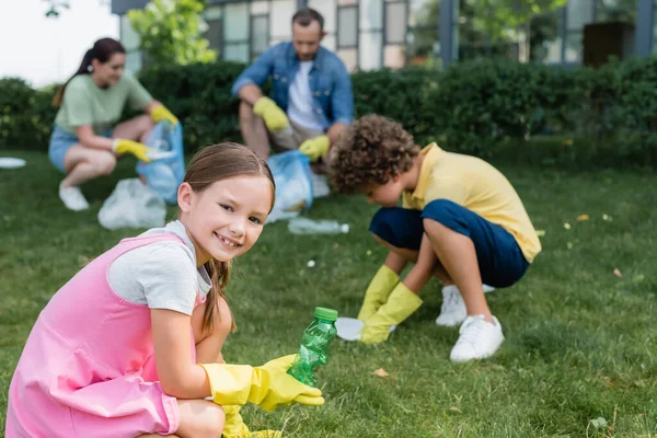 Sonriente chica sosteniendo botella cerca borrosa hermano y padres recogiendo basura al aire libre - foto de stock