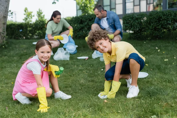 Des enfants souriants dans des gants en caoutchouc tenant des ordures près de la mère et du père brouillés sur la pelouse — Photo de stock