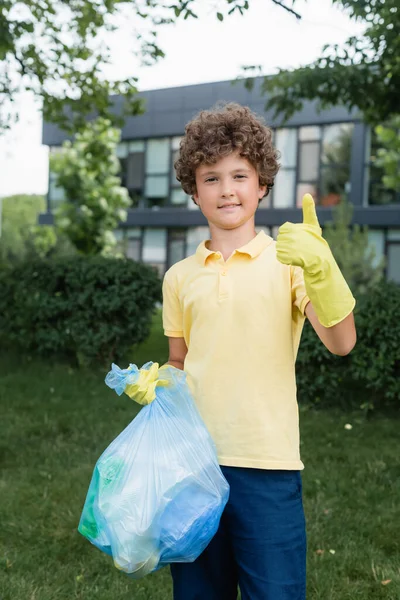 Sonriente niño en guantes de goma sosteniendo la bolsa de basura y mostrando como gesto al aire libre - foto de stock