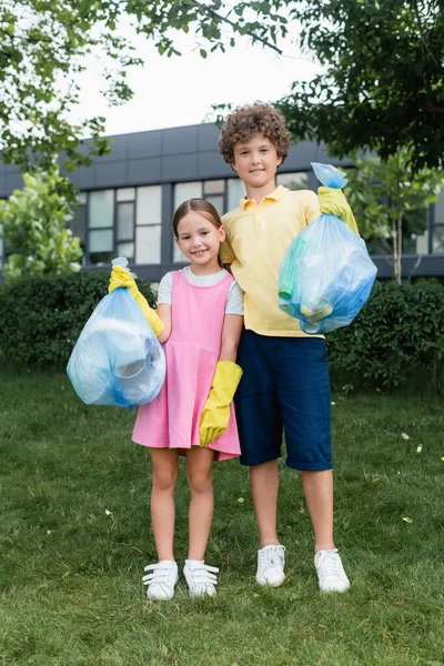 Sonriente chico sosteniendo bolsa de basura cerca de la hermana en el césped - foto de stock