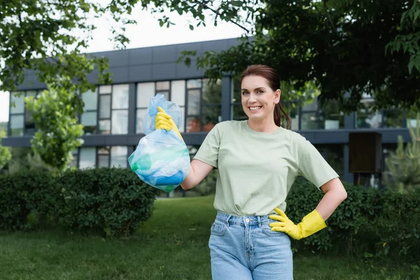 Mujer positiva en guantes de goma sosteniendo bolsa de basura y la mano en la cadera al aire libre - foto de stock