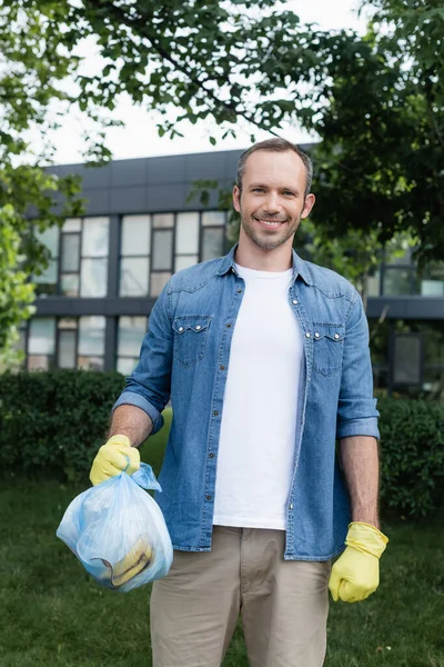 Smiling man in rubber gloves holding bag with garbage and looking at camera outdoors — Stock Photo