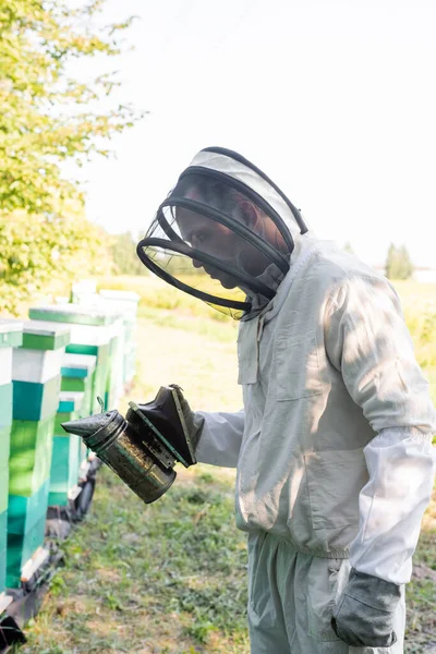 Imker in Schutzanzug und Helm hält Bienenräucher in der Nähe von Bienenstöcken — Stockfoto
